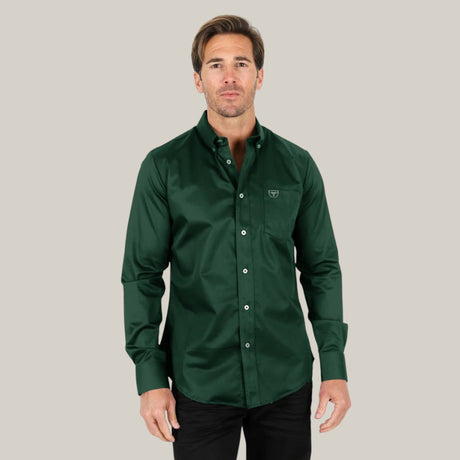 A man in a dark green Mens Single Pocket Logo Modern Fit Stretch Dress Shirt - CTL9667 with long sleeves and black pants stands against a plain beige background, featuring a small logo on the chest pocket as he looks directly at the camera.