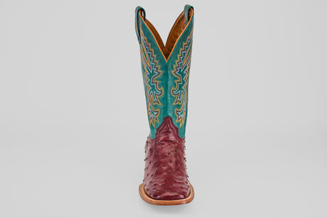 The Rusty - Wine - Square Toe cowboy boot features a wine-hued ostrich leather foot and a tall teal shaft with detailed yellow and white stitching, displayed facing forward on a plain light background.