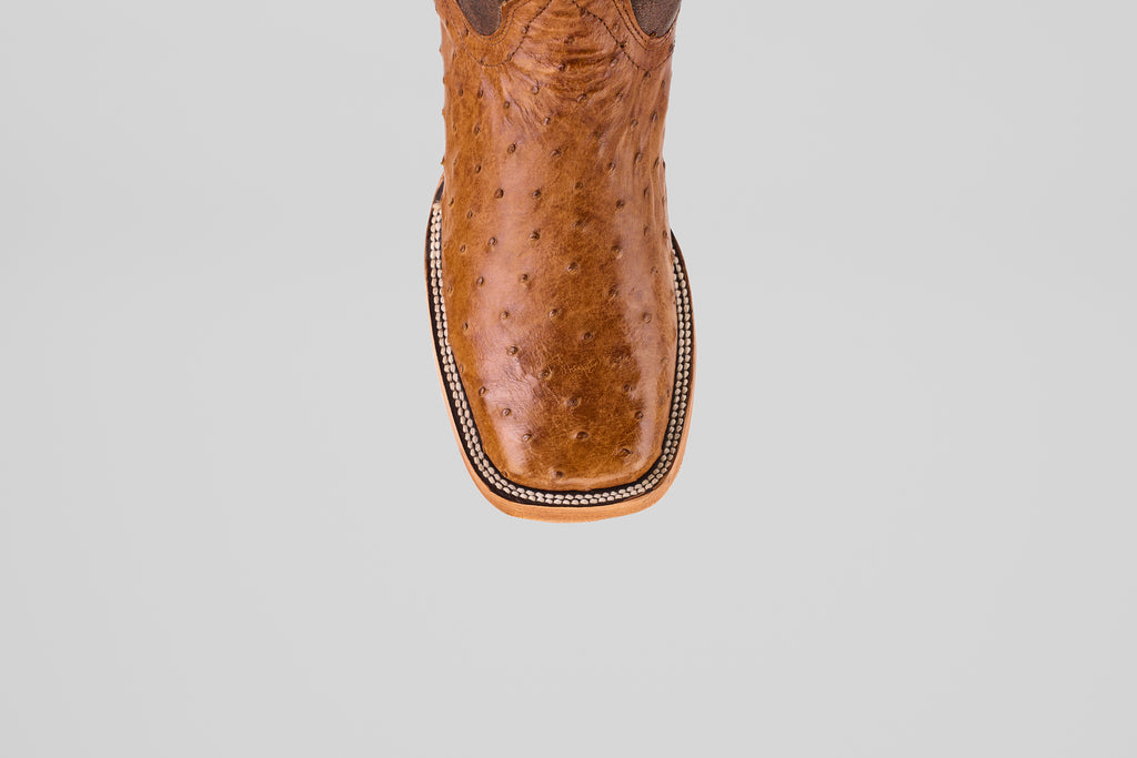 A top view of the Exotic Full Quilt Ostrich - Brandy Brucciato boot features brown leather, a square toe, and decorative edge stitching in classic cowboy style, set against a plain light gray background.