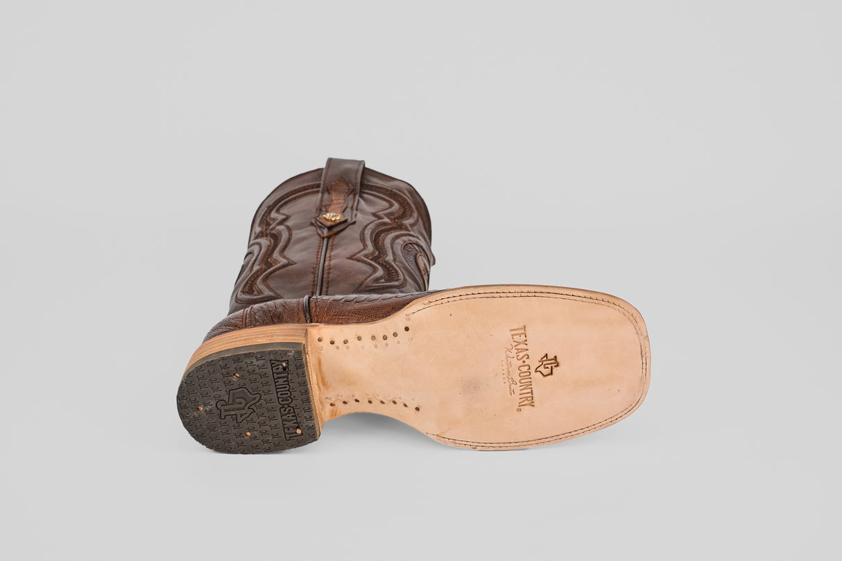 The Austin Leg - Brown - Square Toe cowboy boot lies on its side, displaying a tan sole with black heel, visible stitching, and a Texas Country stamp with Texas icon—showcasing classic western craftsmanship.