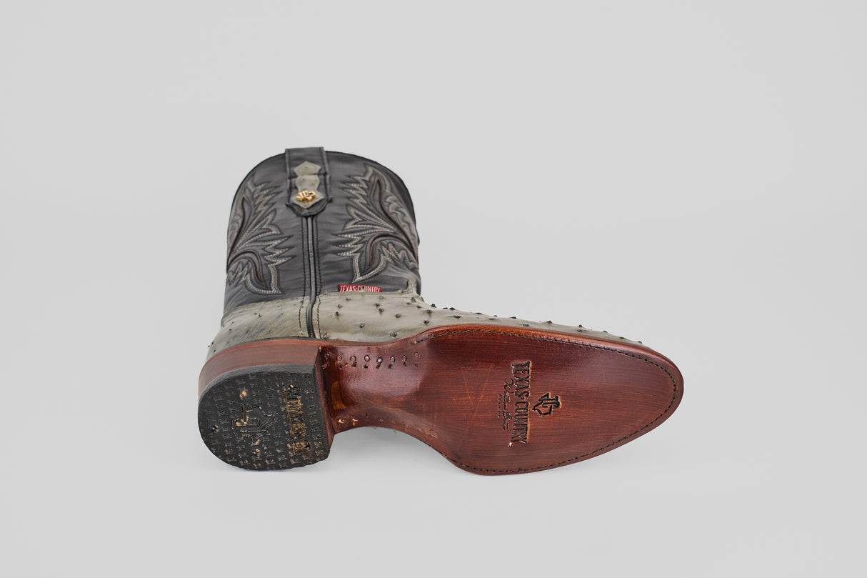 The Austin - Oxford - Round Toe men's cowboy boot is shown on its side, highlighting the stitched upper, grey ostrich leather foot, and a brown sole stamped with Texas Custom Boots. The worn heel rests against a plain light grey background.