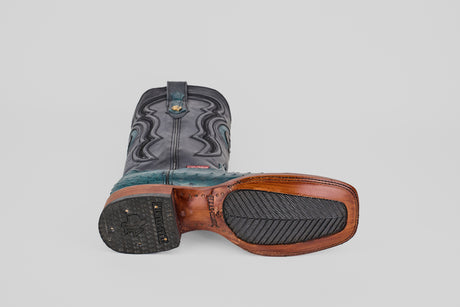 The Austin - Gasoline Green - Square Toe men's western boots feature black, teal, and brown leather. One boot stands upright to display its decorative shaft; the other lies sideways, revealing a textured sole with rubber grip and detailed stitching.