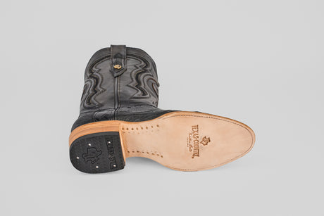 An Austin Leg - Black - Roper Toe boot lies on its side, revealing a tan leather sole stamped with TEXAS COUNTRY and a Texas outline. It features decorative stitching, a stacked leather heel, and the signature roper toe shape.