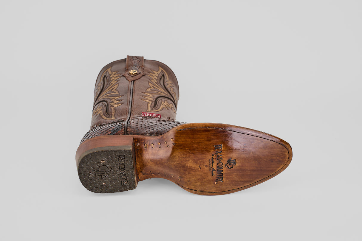 The Tanner - Brown - Round Toe cowboy boot is shown on its side, featuring intricate shaft stitching and a textured, partially worn sole with the Texas Country logo and Python Cafe Oval imprint.