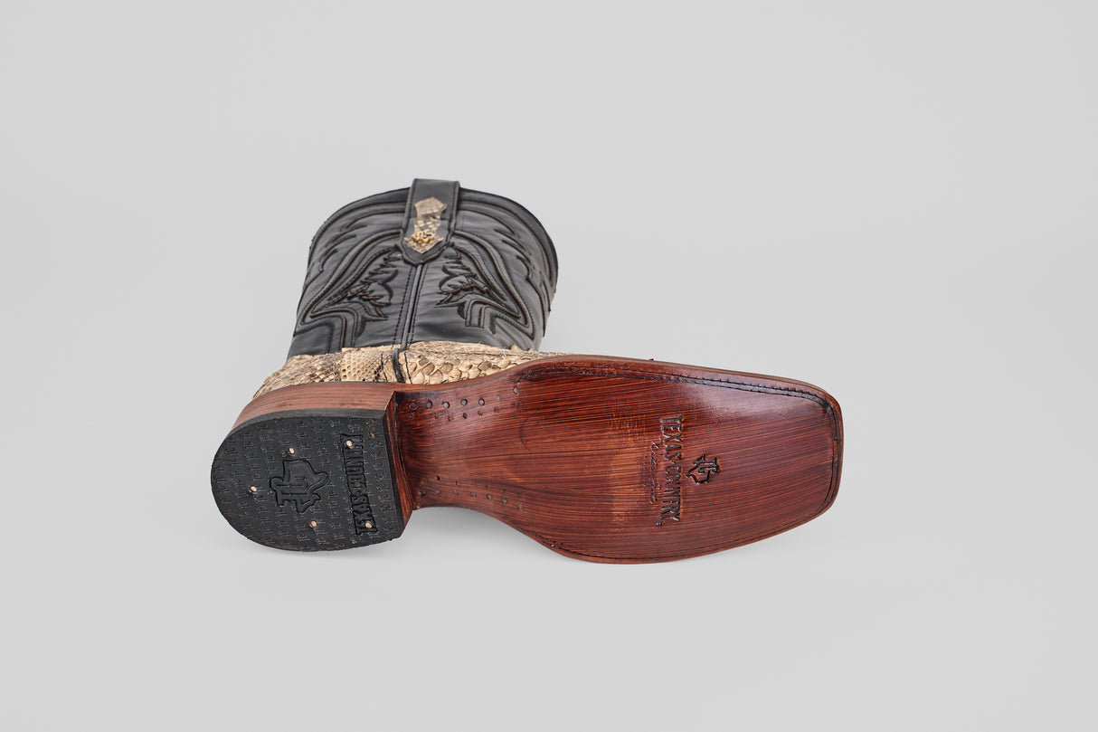 The Tanner - Natural - Rodeo Toe cowboy boot lies on its side, sole up, displaying a wooden heel and detailed engravings. Its leather features striking black and brown patterns over a light gray background.