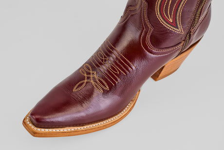 A close-up of the Allison Blackberry - Tall Shaft - Snip Toe boot, featuring a polished dark brown finish, decorative stitching, a pointed snip toe, and a light wooden heel, set against a plain light gray background.