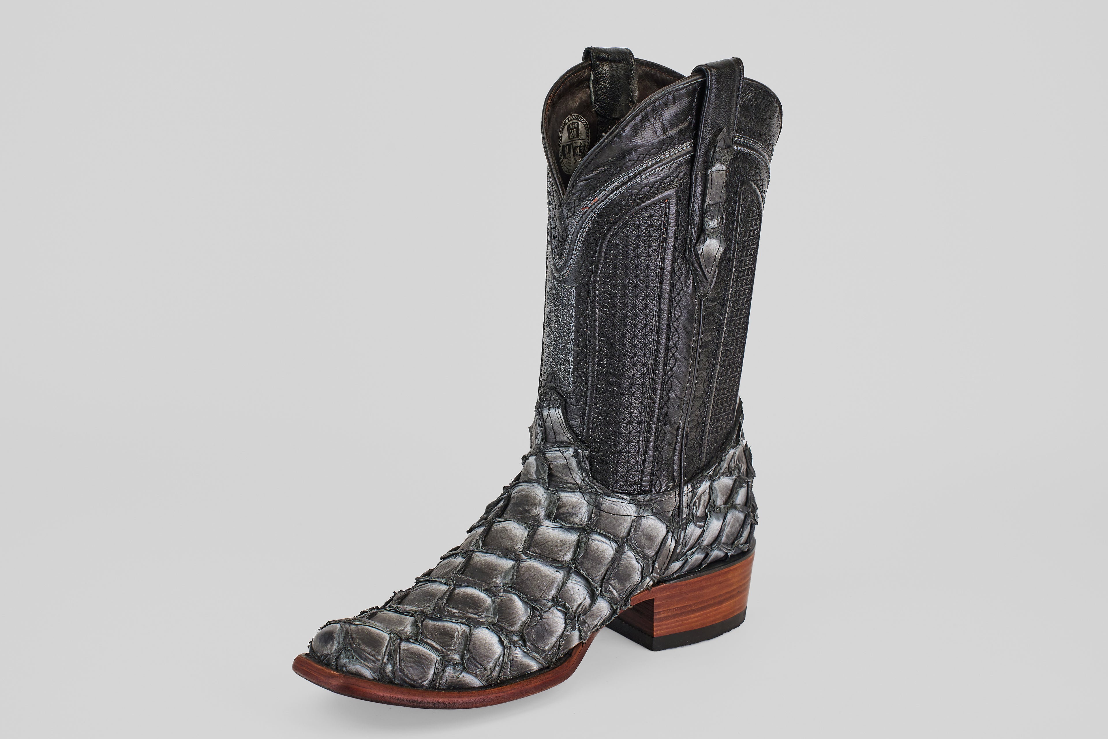 The Conor - Newspaper boot features a black, narrow square toe with a scale-like lower pattern reminiscent of fish leather, a smooth leather upper, pull straps, and a brown wooden heel, all showcased on a white background.
