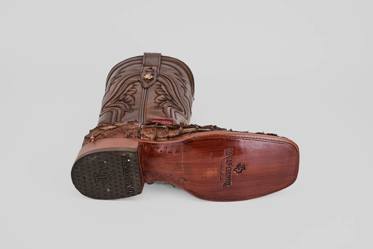 The Conor - Brown - Rodeo Toe cowboy boots are shown with one boot upright and the other on its side, revealing detailed sole stitching, a brand logo, and textured patterns that highlight their classic western style.