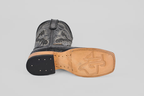 A Caiman Belly Black - Embossed Leather - Square Toe (E423) boot lies on its side, highlighting the tan stitched-logo sole, black heel, and detailed upper stitching against a plain light background.