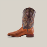 The Cater Honey - Square Toe cowboy boot features a two-tone brown design with intricate stitching, crafted from premium cowhide. The lighter foot and darker shaft are enhanced by a wooden sole and heel, showcasing artisan craftsmanship against a plain white backdrop.