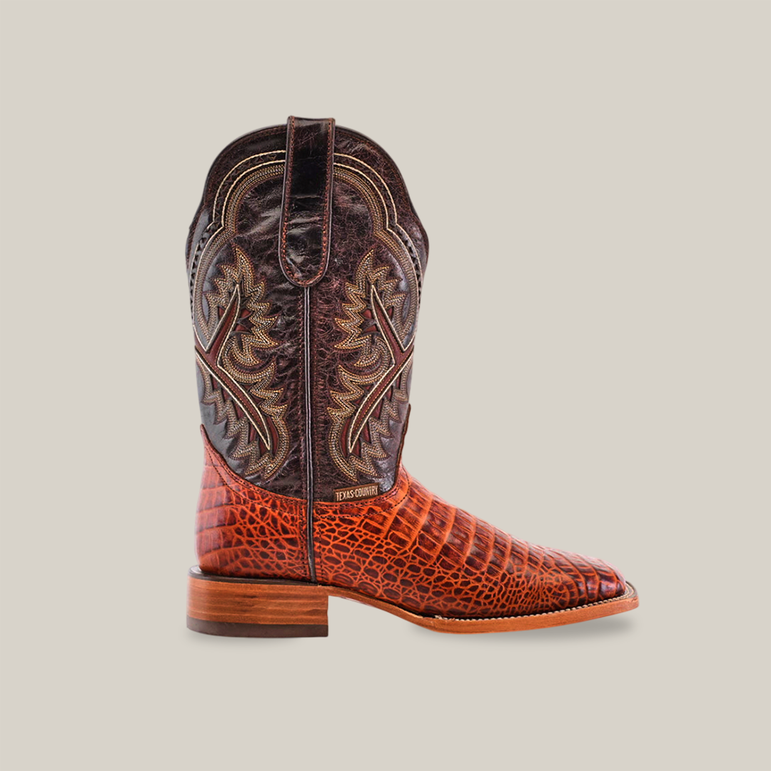 The Caiman Belly Print Cogñac - Square Toe boot features a premium cowhide construction with a brown leather upper, intricate stitching, and a textured reddish-brown foot. It boasts a cognac square toe design paired with a wooden heel on a pristine white backdrop.