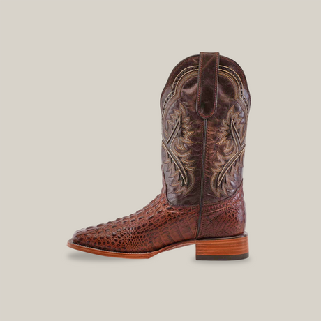 A brown cowboy boot with Caiman Hornback print and intricate stitching is shown on a white background. This artisan-made boot, named Caiman Hornback Print Chocolate - Square Toe, features a low wooden heel, square toe, and top pull strap.