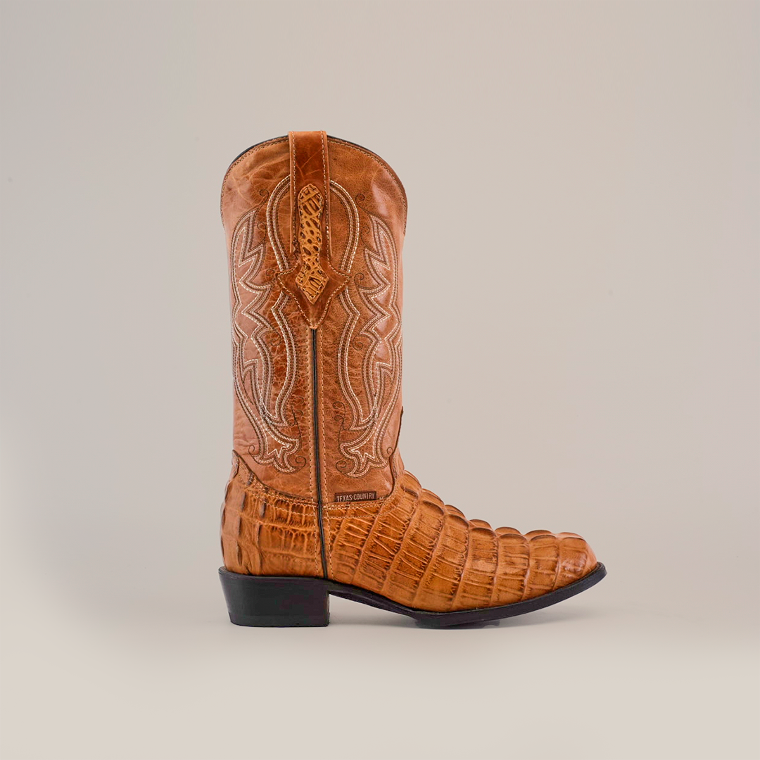 The Crocodile Tail Print Orix - Round Toe features a handcrafted brown cowboy boot with premium cowhide leather, intricate stitching, and a crocodile tail print on the foot. Positioned laterally against a white background, it highlights a black heel and decorative pull strap.