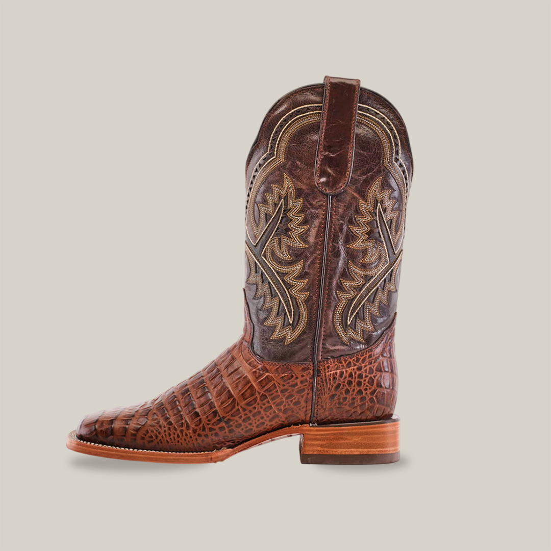 The Caiman Belly Print Chocolate features a handmade cowboy boot with textured leather and intricate stitching. It has a square toe, low stacked heel, and is displayed against a plain white background.