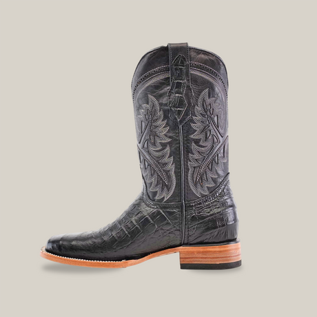 The Caiman Belly Print Black - Square Toe cowboy boot, crafted from premium cowhide leather, showcases intricate stitching and a textured surface. It features a wooden sole and is positioned sideways on a plain white background, emphasizing its elegant design.