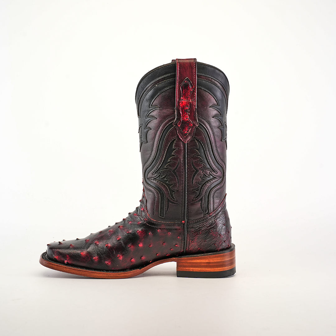 The Exotic Ostrich - Black Cherry cowboy boot, crafted from genuine ostrich leather, features a wooden heel and intricate red-accented patterns. Its textured surface and ornamental detailing with a stylish rodeo toe stand out against a plain white background.