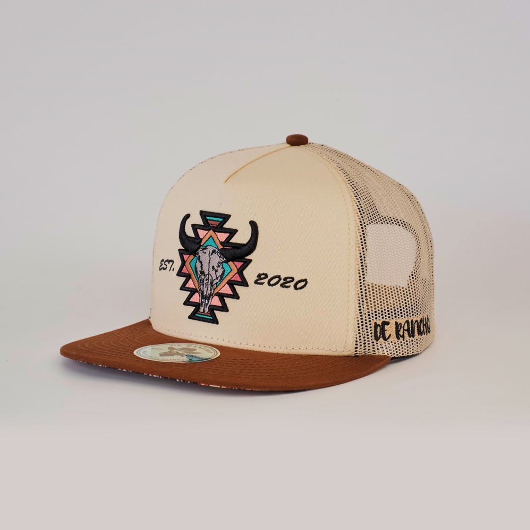 The De Rancho Cap Aztec Camel is a tan and brown baseball cap with a stylized bull skull and geometric shapes, celebrating Western heritage. Featuring Est. 2020 and De Ranch in black embroidery, it has a solid front panel and mesh back that showcase ranch life-inspired embroidered designs.