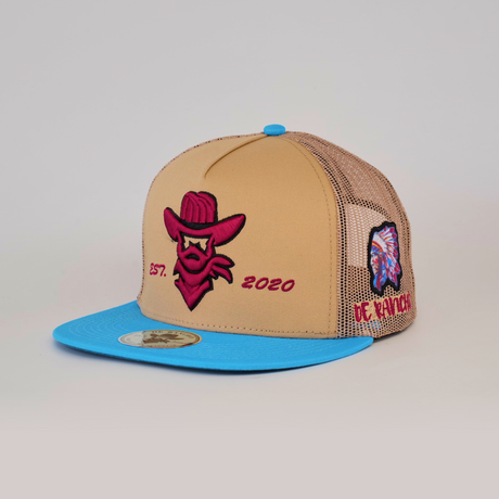 The De Rancho Cap Classic Camel is a tan and blue trucker hat featuring a red cowboy logo with EST. 2020, celebrating Western heritage. The side boasts an embroidered Native American chief and OE RANCH text, along with a gold sticker on the brims underside.