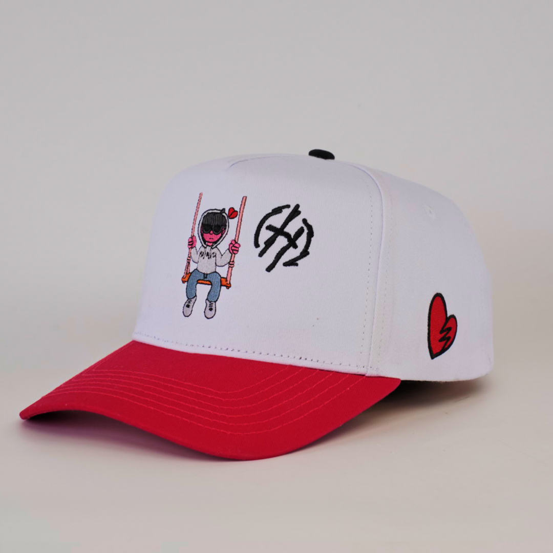 The De Rancho Cap Jr H White is a white cap with a red brim, featuring an embroidered person on a swing with a bear mask. It embraces Western heritage with a black logo on the front and a small black and red broken heart design on the side.