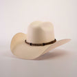 The 5000x Chihuahua Straw Hat, featuring a cream color, wide brim, and brown band, showcases exquisite craftsmanship as it sits angled on a white surface.