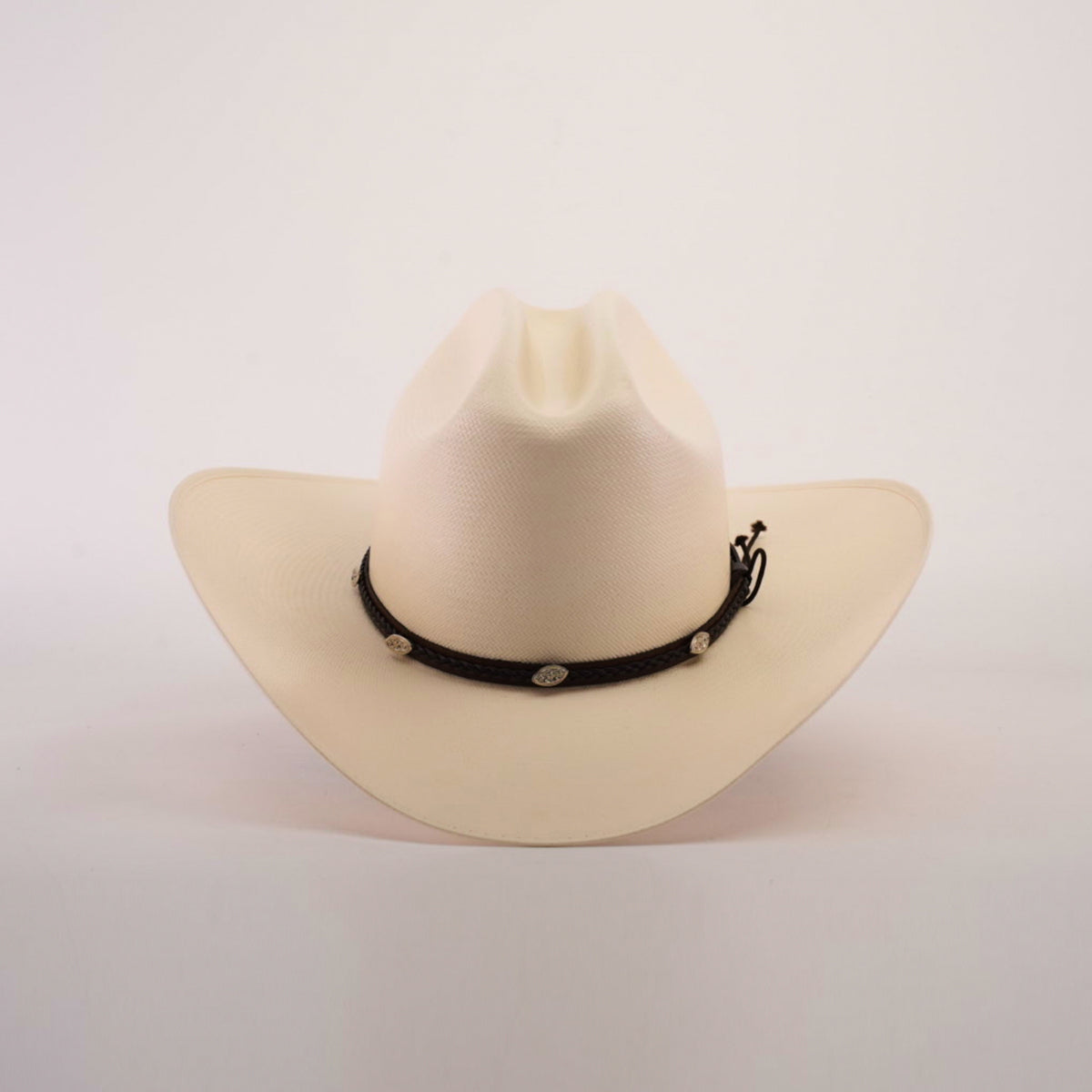 The 1000X Sinaloa Straw Hat features an elegant design with a wide brim, a black band adorned with silver conchos, and a plain white background.