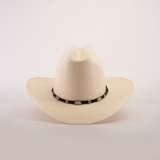 The 1000X Dos Carnales Straw Hat, an exceptional beige cowboy hat with a black band and decorative white oval accents, is displayed against a plain white background.