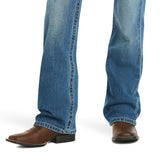 A young boy stands with legs slightly apart, wearing Ariats medium wash boot cut jeans with a faded look and brown leather cowboy boots featuring a pointed toe and block heel, set against a white background.