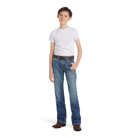 A young boy stands confidently in a relaxed pose wearing a white t-shirt, Ariat Boys B4 Boundary Dakota Medium Wash Relaxed Fit Boot Cut Jeans (10018345), a brown belt, and brown shoes against a plain white background.