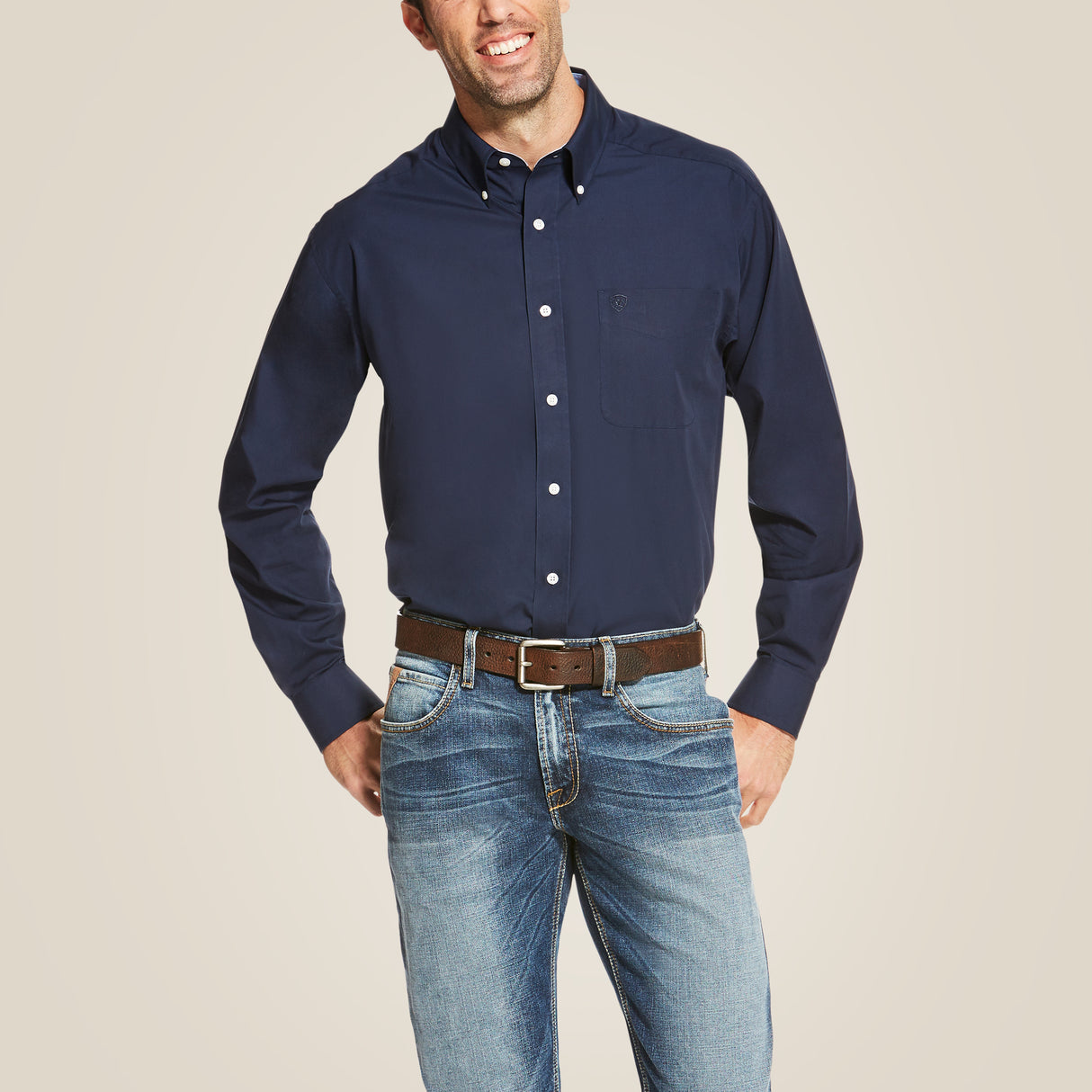 A man in a Wrinkle Free Solid Classic Fit Shirt Navy - 10020330 and blue jeans stands with a hand in his pocket and the other on his hip. With a brown belt, he smiles against a plain background, highlighting the shirts wrinkle-resistant marvel.