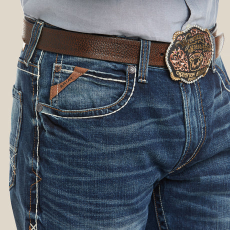 A close-up of someone in distressed blue jeans, identified as Ariats M4 Low Rise Stretch Preston Boot Cut Jeans in Silverton, paired with a light gray shirt. Theyre wearing a dark brown leather belt with an ornate gold buckle. The jeans feature contrast stitching and visible brand tags.