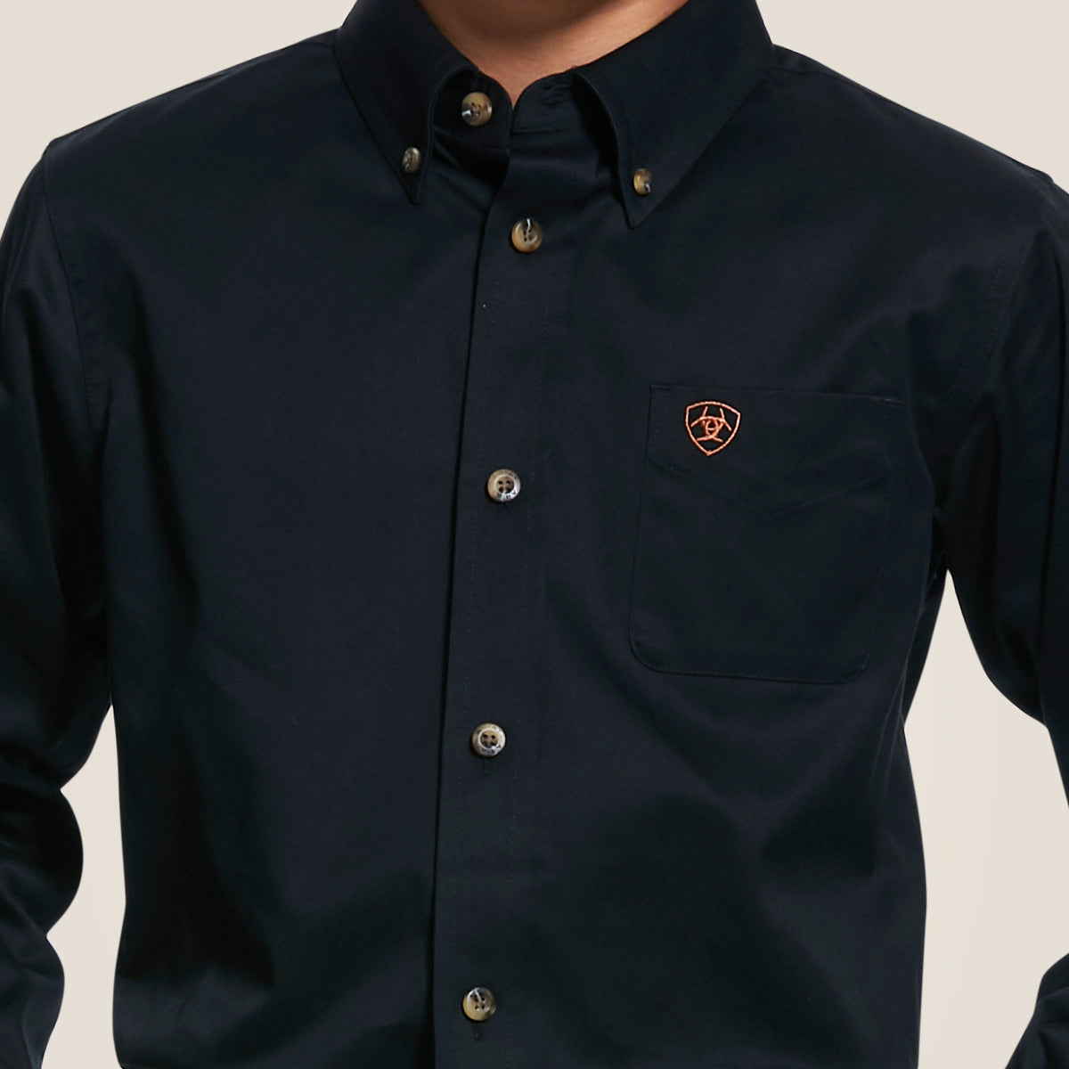 A kids wears the Ariat Black Solid Twill Classic Fit shirt, featuring a left chest pocket with a red and gold shield emblem. The collared black button-up shirt stands out against a light beige background.