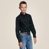 A young person smiles against a plain beige backdrop, wearing an Ariat Black Solid Twill Classic Fit - Kids Boys Shirt (10030161) with a logo on the chest and blue jeans.