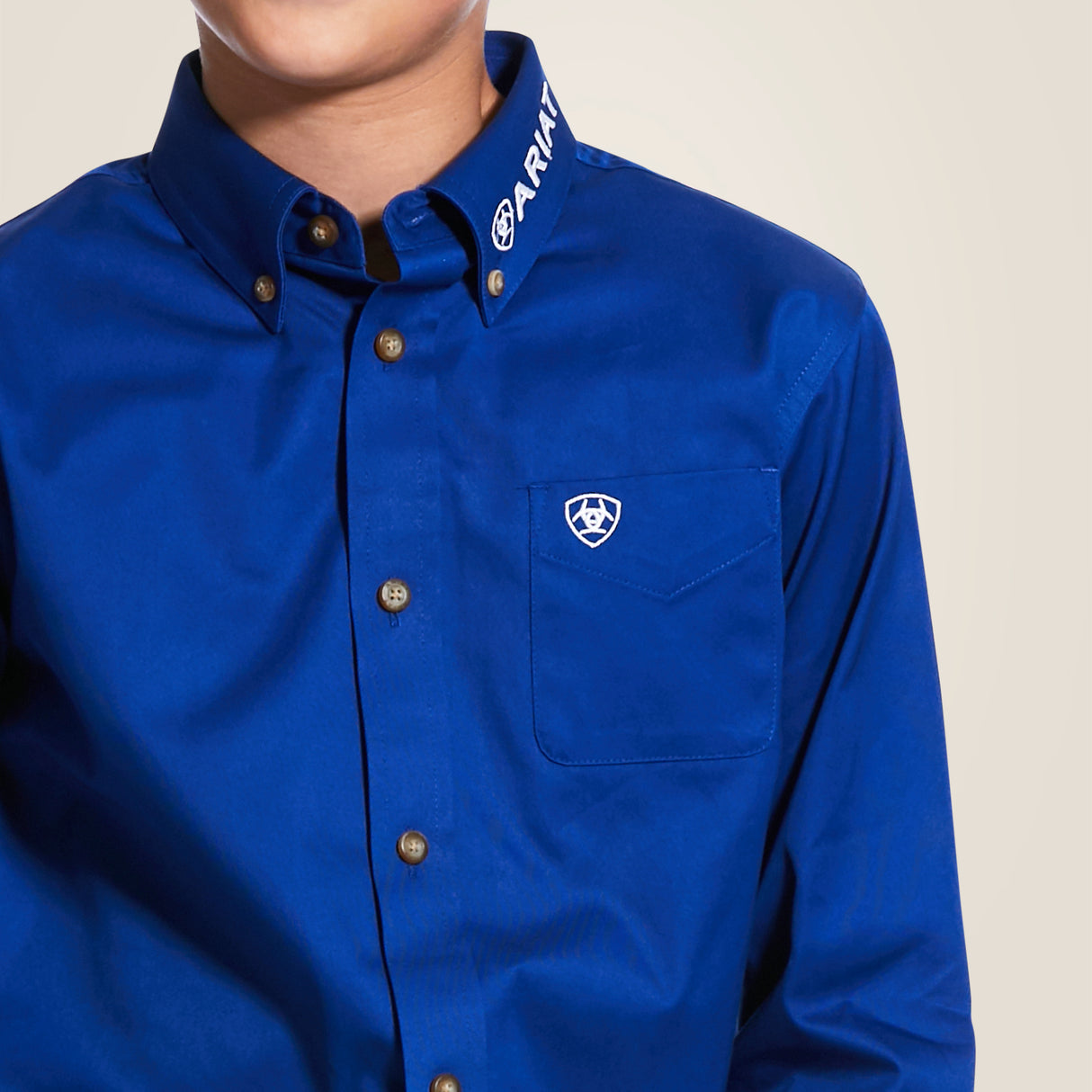 A person wears the Ariat Boys Blue Team Logo Long Sleeve Western Shirt, featuring a durable cotton-blend twill, a logo pocket, and ARIAT on the collar. Their face is not visible.