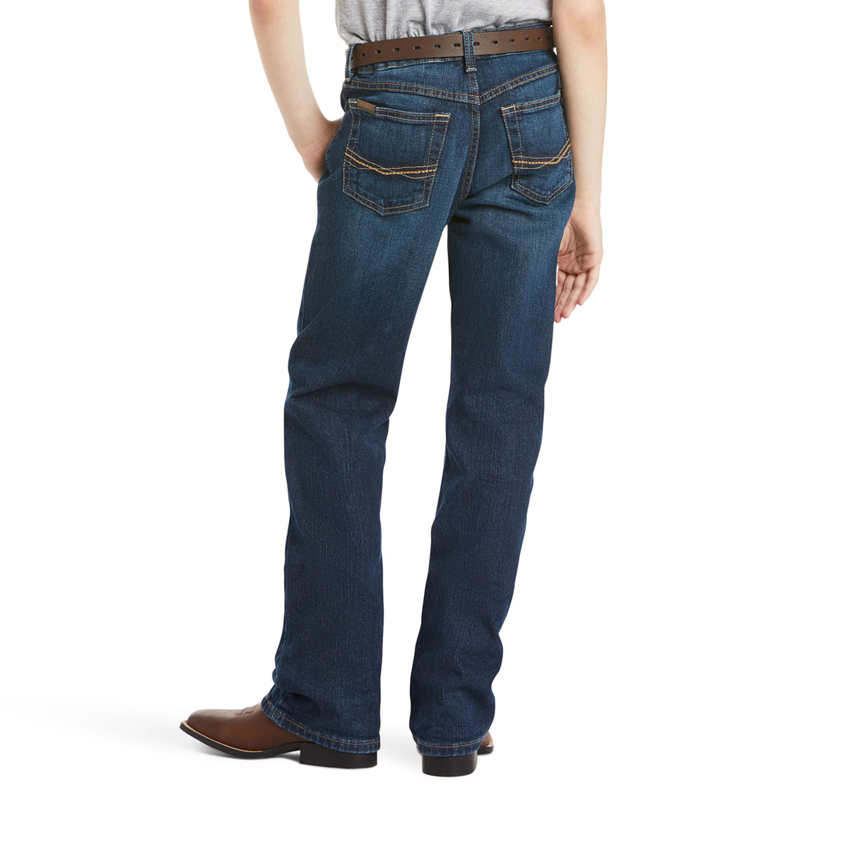 Rear view of a person in Ariat Boys B4 Legacy Dark Wash Relaxed Fit Boot Cut Jeans with visible stitching, paired with a gray shirt, brown belt, and cowboy boots. Their left hand rests in the back pocket.