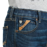 A person is wearing Ariat Boys B4 Legacy Dark Wash Relaxed Fit Boot Cut Jeans, featuring yellow stitching, a small front pocket with a brown leather Ariat label, and a silver button on the waistband. A gray shirt peeks out above the jeans.