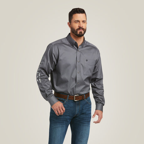 A man with short dark hair and a beard is wearing the Team Logo Twill Classic Fit Shirt in gray, which features embroidered details on the right sleeve. He pairs this with blue jeans and a brown belt against a plain, light gray background.