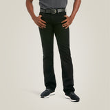 A person stands against a plain background, wearing a gray shirt, black belt, M7 Slim Legacy Straight Jeans in black (10037890), and sleek black sneakers with white soles. Their hands rest confidently on their hips in a modern cowboy style. The photo is cropped from the neck down.