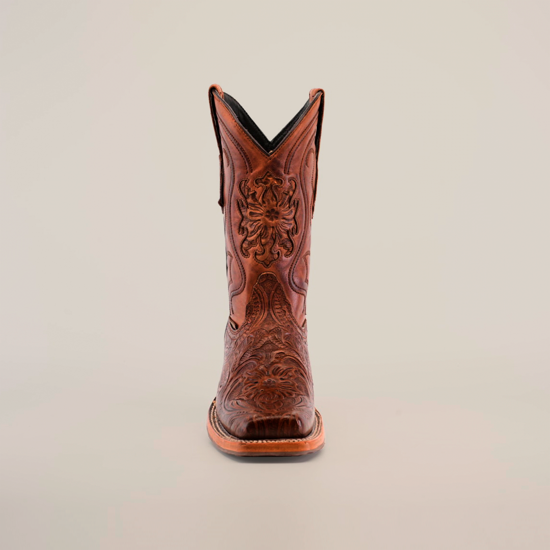 The Handtooled Leave Brown boot, featuring a Rodeo Toe, is handcrafted from premium cowhide with floral and geometric designs. It boasts a slip-resistant sole, pointed toe, and slightly raised heel against a plain light background.