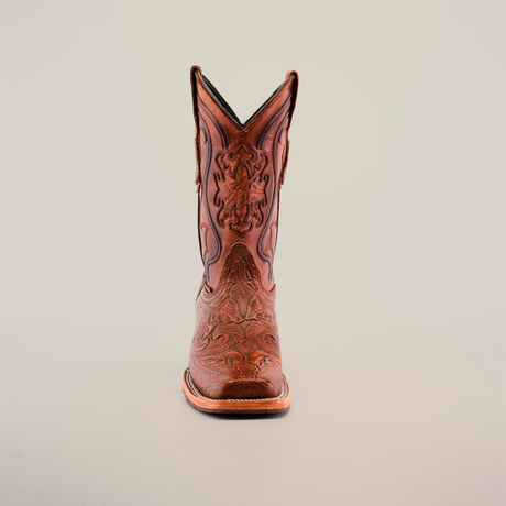 A single handcrafted brown cowboy boot, the Handtooled Leave Cogñac, stands on a white background. Made from premium cowhide leather, it features intricate patterns and stitching with a rodeo toe design that highlights its artisanal craftsmanship.