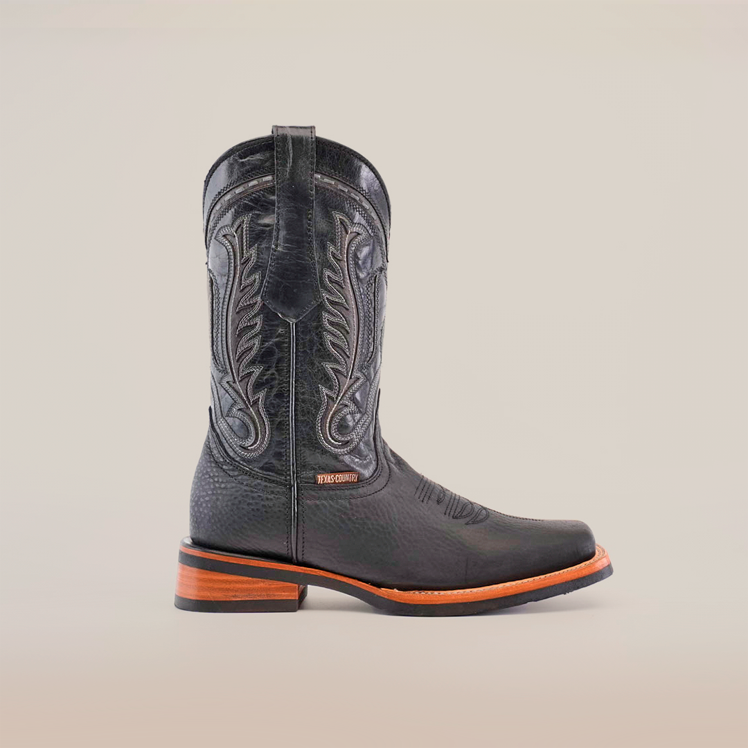 The Cheyenne Black cowboy boot boasts detailed shaft stitching, premium cowhide leather, a brown wooden heel, and a rodeo toe. Its slightly pebbled texture and slip-resistant sole enhance its rugged appeal against a plain white background.