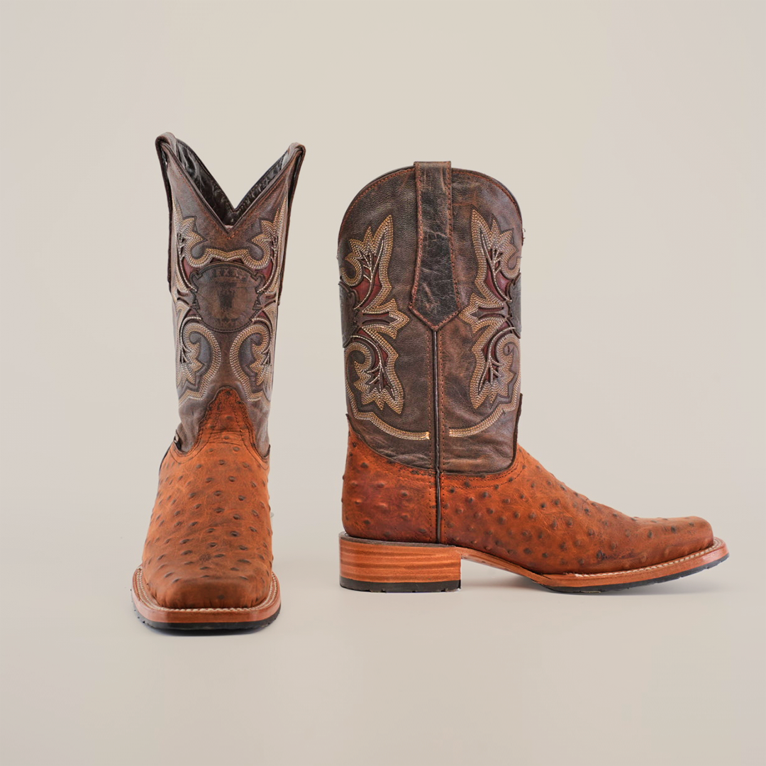 A pair of Karoo Ostrich Roughout Print Cogñac boots, showcasing intricate shaft stitching and an ostrich print foot, features a slip-resistant sole. The primarily brown cowhide design with darker hues is set against a plain white background.