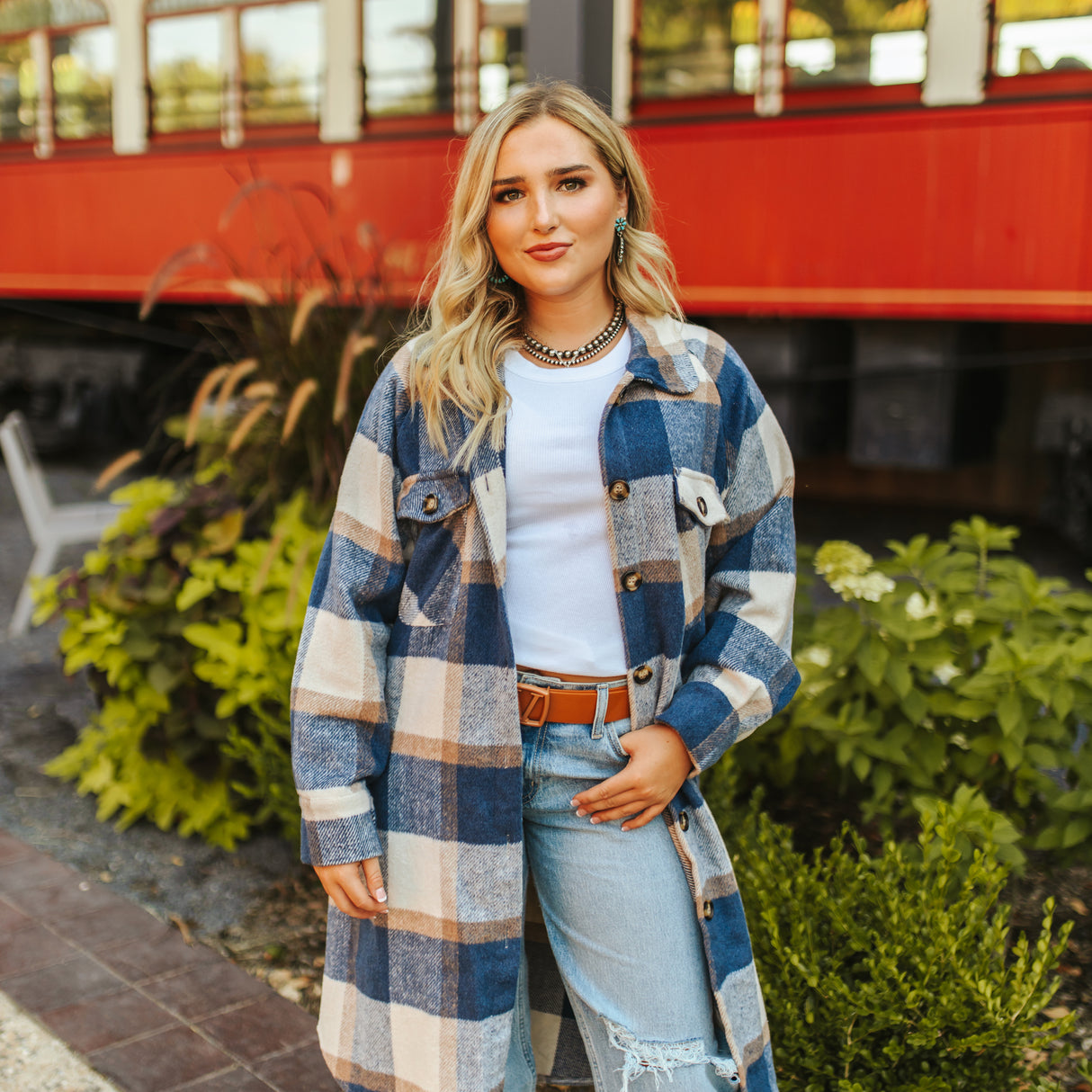 A person with long blonde hair stands in front of a red vintage train car, exuding a cozy, stylish vibe. Theyre wearing the LB Blue Plaid Fleece Long Button Down JK223-BL coat, with a white top, ripped jeans, belt, and necklace. Green foliage adds charm to the scene.