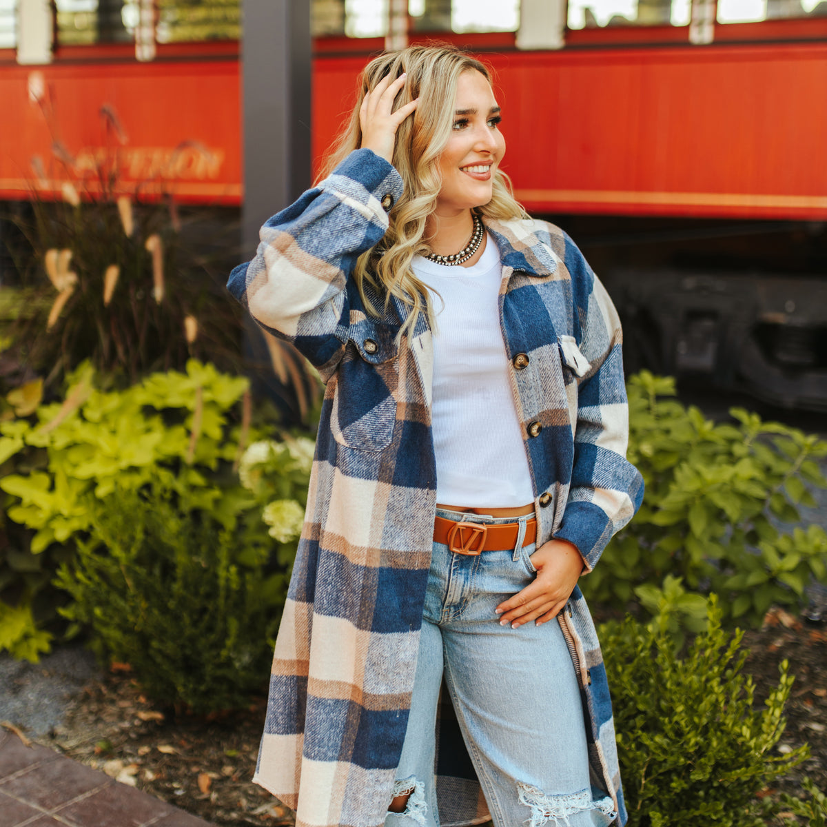 Dressed in the LB Blue Plaid Fleece Long Button Down JK223-BL with ripped jeans, a white shirt, and a brown belt, a person stands smiling in front of a red train and leafy plants. Hand in hair, they epitomize womens chilly day essentials.