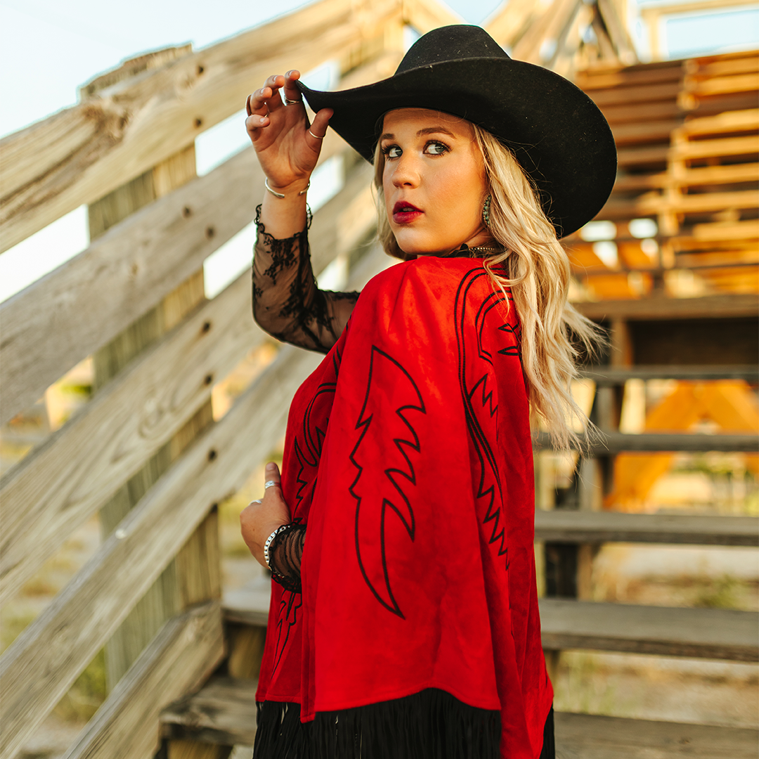 A woman in a black cowboy hat and the LB Red Suede Stitch Cape J0059-Red with black designs stands on a wooden staircase. Her striking draped silhouette, with hand on her hat, is enhanced by the bright sky. Her fringed cape perfectly complements her lace sleeves.