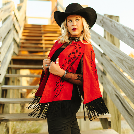 A woman stands on a wooden staircase in a black cowboy hat and the LB Red Suede Stitch Cape J0059-Red, her silhouette catching the eye. She gazes to the side, wearing black lace and silver rings. The outdoor setting features warm lighting and rustic charm.