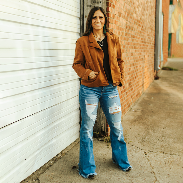 A woman smiles while standing against a white garage door next to a brick wall, giving off an urban yet rustic vibe. She wears an LB Camel Suede Western Stitch Blazer with ripped blue jeans and black shoes—a seamless mix of modern style and timeless elegance.