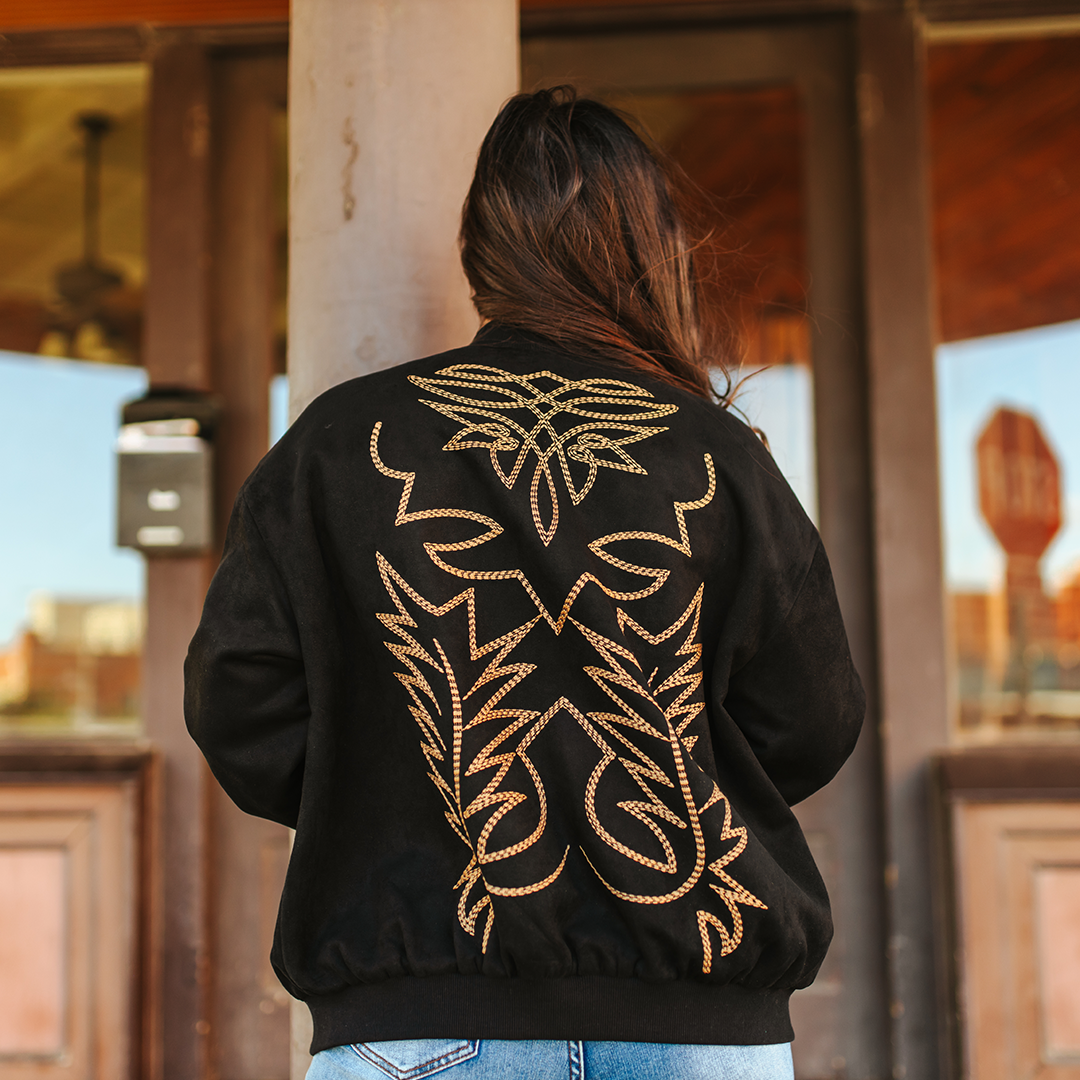 With long hair, a person stands with their back to the camera wearing the LB Black Western Stitch Bomber JO241-BLK. The jacket features intricate gold embroidery, exuding edgy elegance as they pose in front of a wooden building with large windows and a visible fan inside.