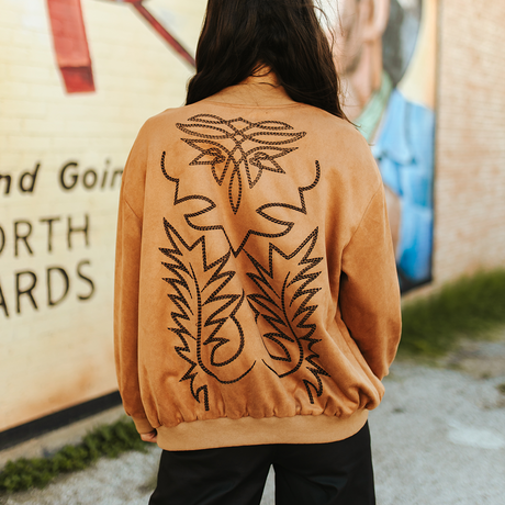 A modern woman with long dark hair stands with her back to the camera, wearing the LB Camel Western Stitch Bomber JO241-CML, showcasing its black embroidered Western details. She exudes effortless style near a mural on a brick wall.