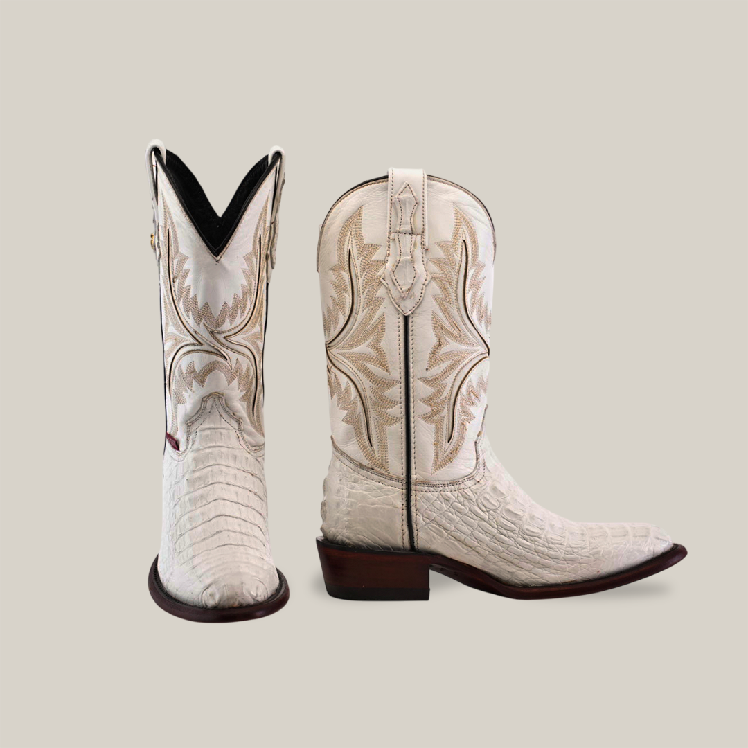 The Exotic Caiman Hornback in white are handmade boots crafted from genuine Caiman leather, featuring intricate tan stitching on crocodile-textured shafts. One upright boot highlights its wooden heel and pointed toe against a beige background.