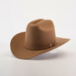 The 6X 2 Carnales Fawn is a brown felt cowboy hat with a classic curved brim, top crease, and decorative band featuring a metal buckle, all set against a neutral background. This iconic piece embodies timeless western style and rugged elegance.